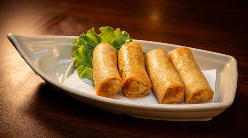 Four golden-brown spring rolls on a white boat-shaped plate with a lettuce leaf.